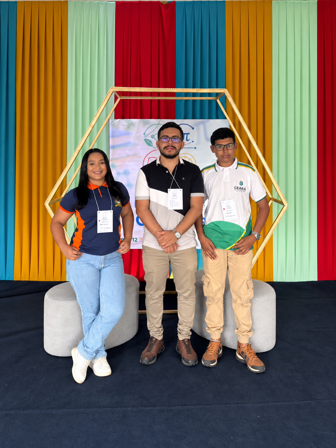 Três pessoas com crachás posam diante de um painel e cortinas coloridas, em registro relacionado ao projeto VistoTECH.